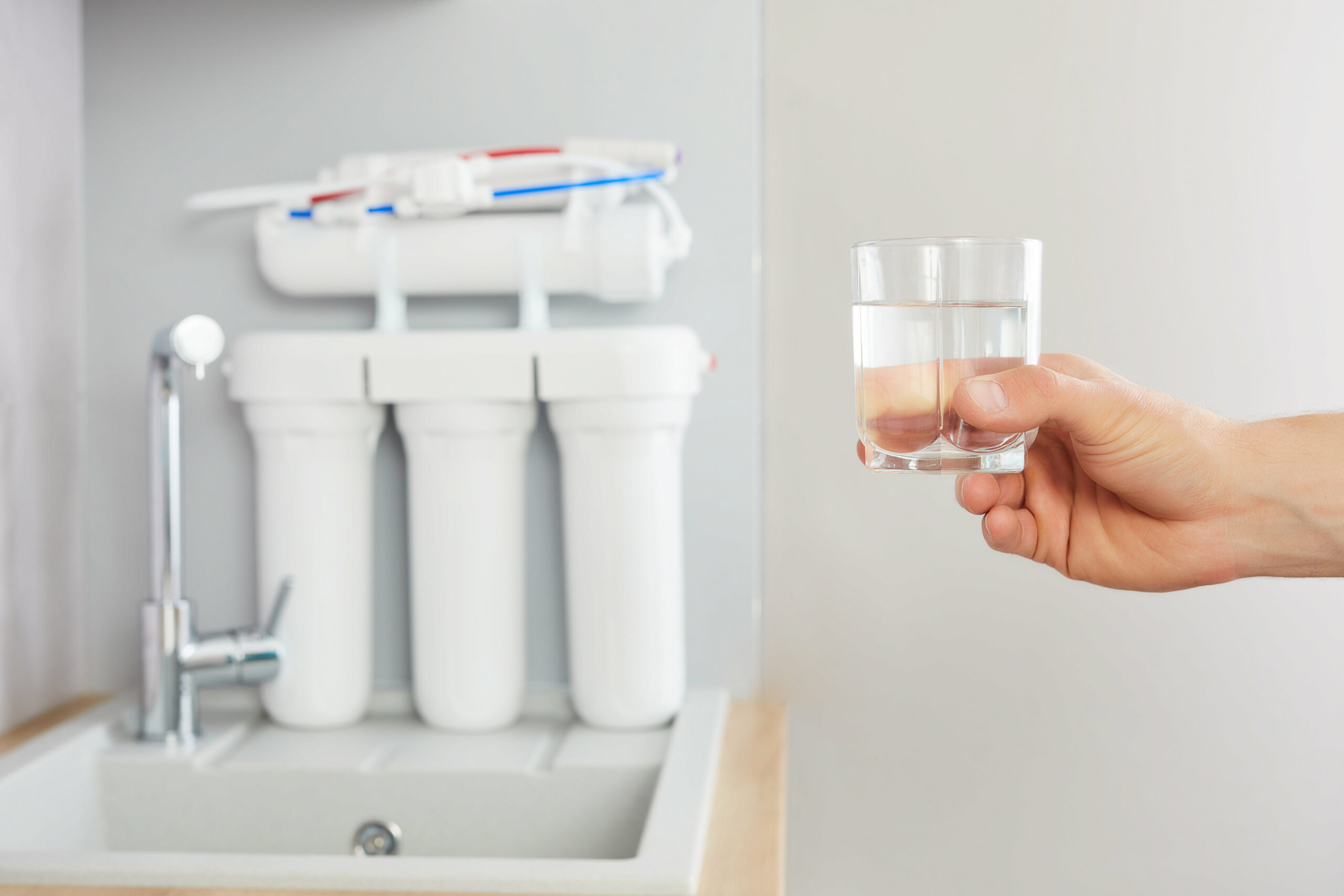Men’s hand holds a glass of clear water. Tap and reverse osmosis filter in the background.